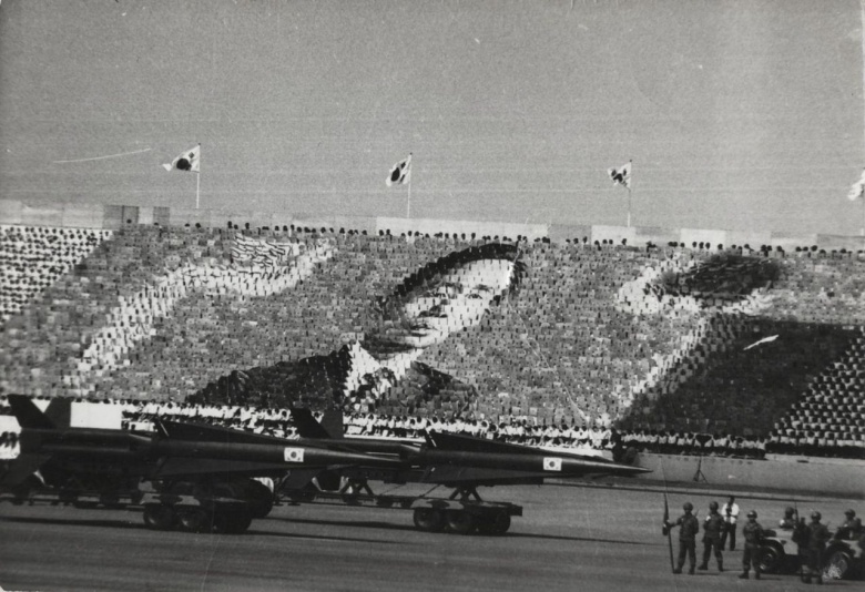 Прижизненный культ личности Пак Чон Хи в Южной Корее 1970-х: зрители военного парада держат карточки, складывающиеся в портрет диктатора. Сеул, 1 октября 1973 года