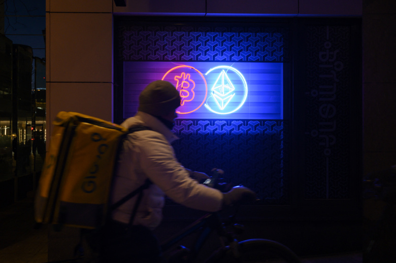 Курьер доставки еды проезжает мимо неоновой вывески, рекламирующей криптовалютную биржу.